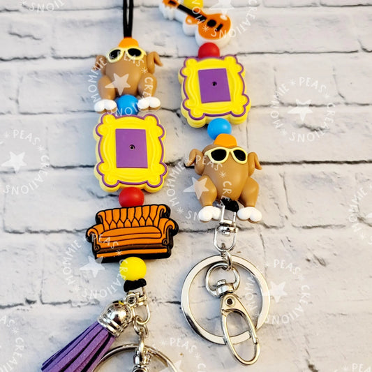 Handmade keychains with yellow and purple frames, brown turkeys with sunglasses, orange sofa charms, and colorful beads on a light brick background, unique gift from Mommie Peas Creations.