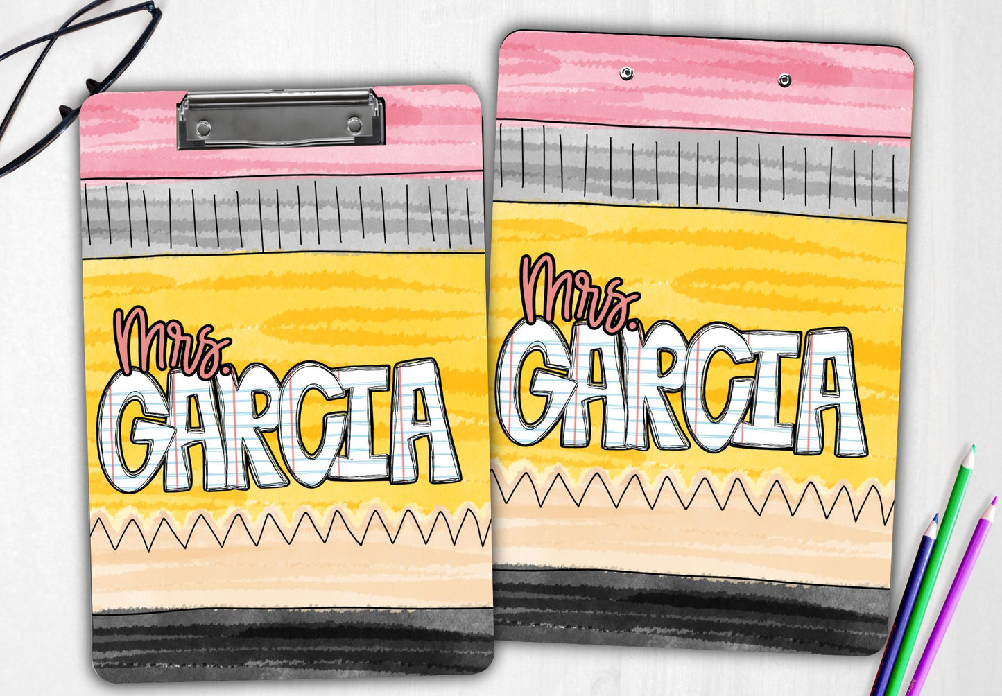 A personalized teacher clipboard featuring a vibrant pencil-themed design with the name "Mrs. Garcia" displayed in large, paper-style letters. The clipboard is shown from both the front and back, highlighting the same playful and colorful design on each side. The background mimics a classic yellow pencil with pink, gray, and black accents. Glasses and colored pencils add a touch of creativity to the scene, making it perfect for teachers or educators.
