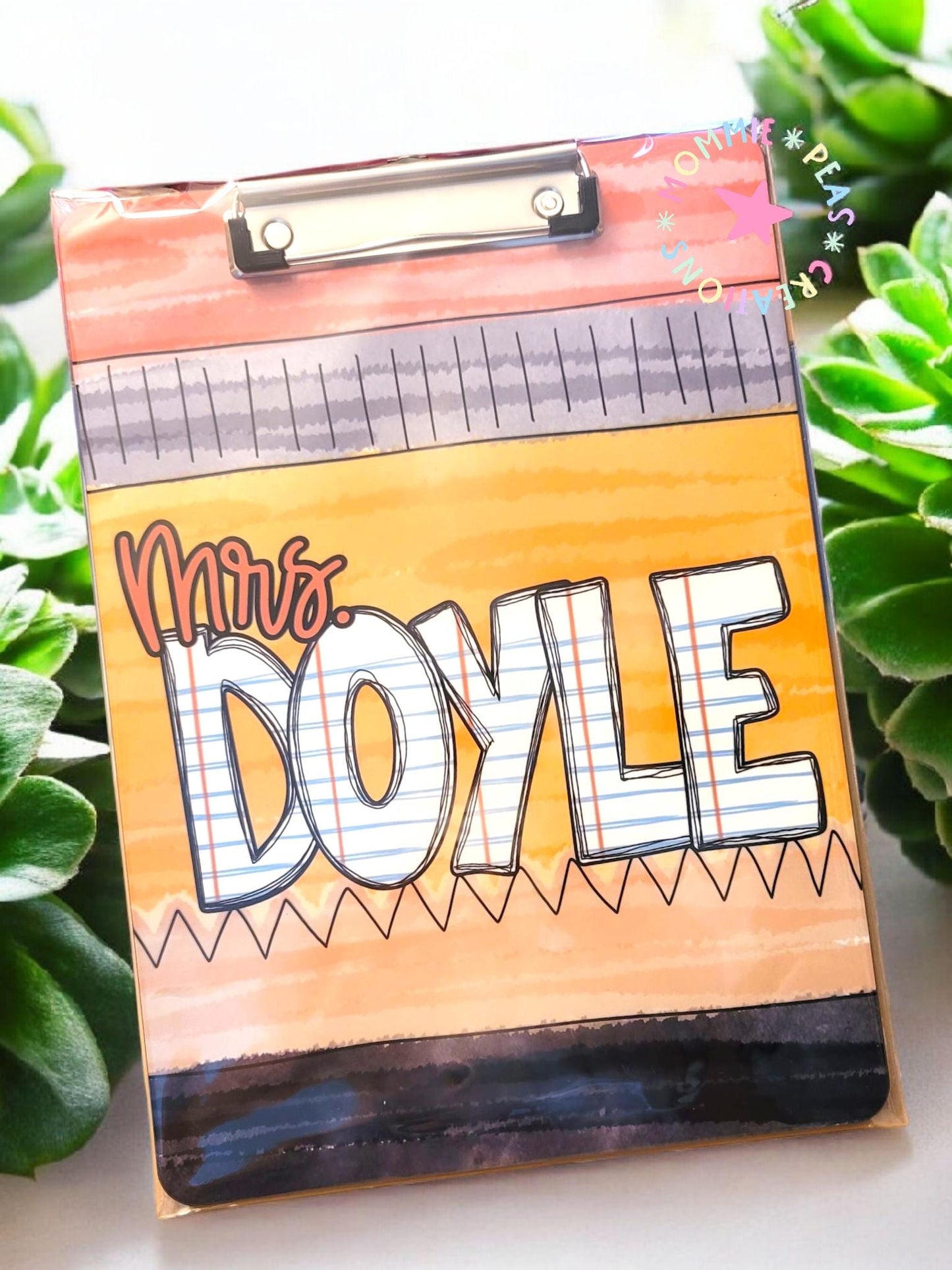 A personalized clipboard featuring the name "Mrs. Doyle" in a fun, school-themed design. The top section is colored in shades of pink and gray, resembling a pencil. The middle section has a yellow-orange background with a zigzag pattern at the bottom. "Mrs." is written in cursive pink, while "DOYLE" is in bold, block letters with a lined notebook paper pattern. A metal clip is visible at the top, and the background includes green succulent plants.