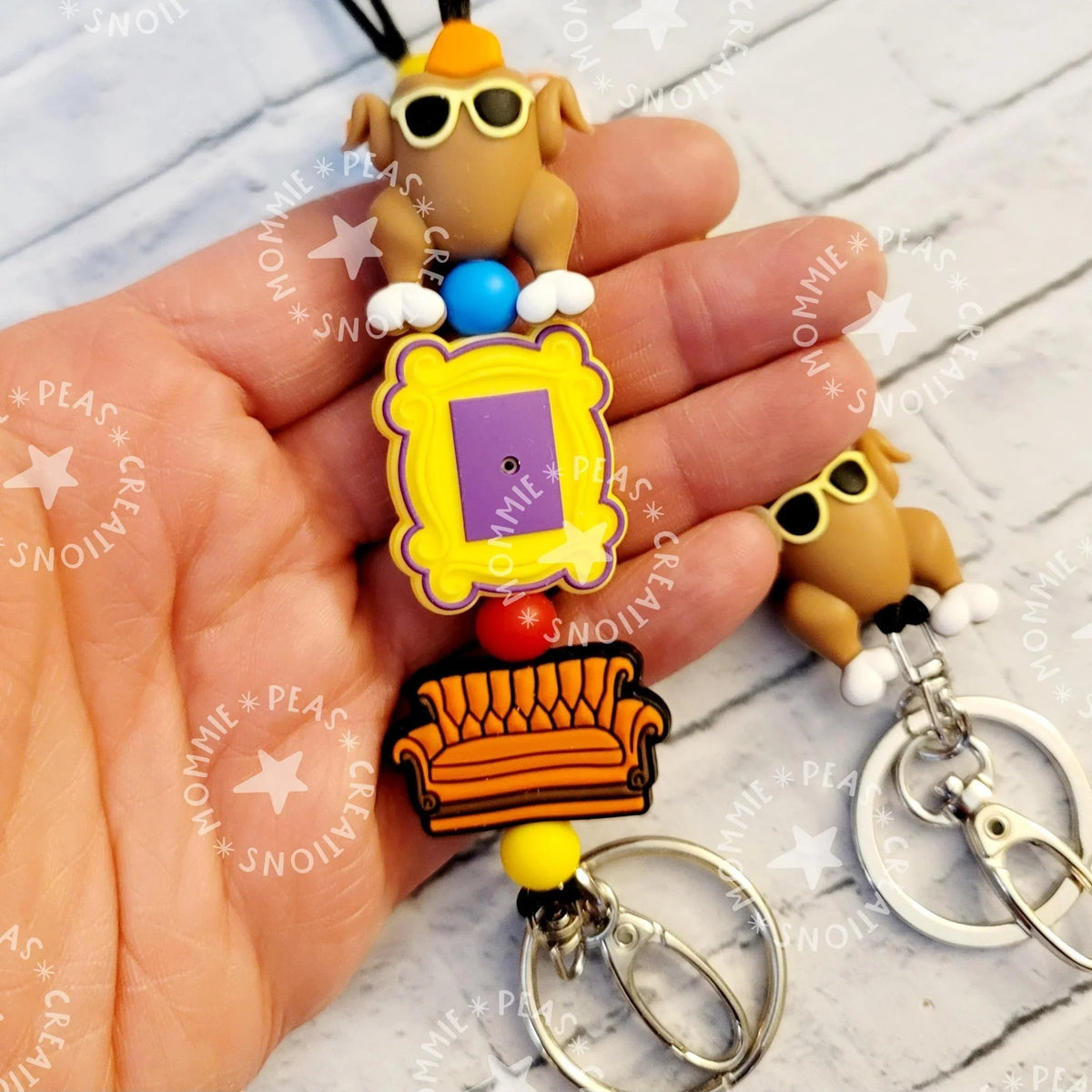 Mommie Peas Creations Fall Turkey Lanyard with Friends-themed charms: a turkey wearing sunglasses, a yellow picture frame, and an orange couch. Features silicone beads and silver hardware.