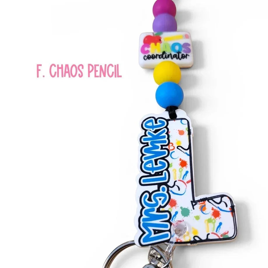 Mommie Peas Creations custom acrylic lanyard with 'Chaos Coordinator' charm and personalized 'Mrs. Lemke' letter 'L' design, featuring colorful paint splatters and school icons.
