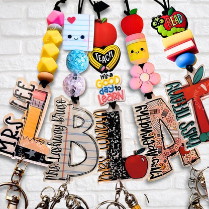 A collection of colorful personalized teacher-themed lanyards with various acrylic monogram letters, vibrant silicone beads, and fun educational-themed charms. The lanyards feature designs such as apples, pencils, composition notebooks, and inspirational quotes like 'Teach, it's a good day to learn.' Some monogram letters have patterns like ruler markings, lined paper, and Composition Design.