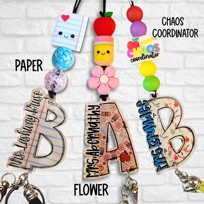Close-up of three custom teacher lanyards with paper, flower, and chaos coordinator themed toppers and acrylic monogram letters personalized with teacher names.