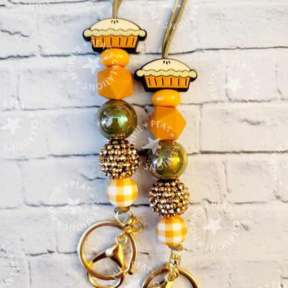 Pair of fall-themed keychains with pie charms, yellow beads, and rhinestone accents on white brick background