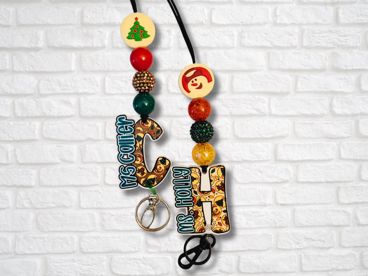 Two festive cookie letter lanyards with personalized charms “Ms. Collier” and “Ms. Holly” from Mommie Peas Creations. They are displayed against a white brick wall, featuring snowman and Christmas tree silicone beads, rhinestone accent beads, and matching paracord.