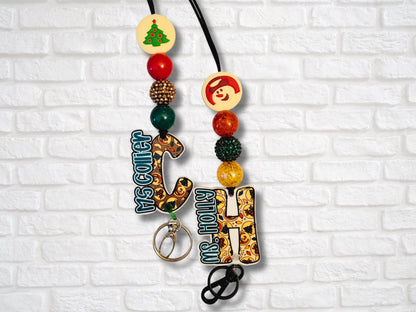 Two festive cookie letter lanyards with personalized charms “Ms. Collier” and “Ms. Holly” from Mommie Peas Creations. They are displayed against a white brick wall, featuring snowman and Christmas tree silicone beads, rhinestone accent beads, and matching paracord.