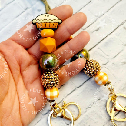 Hand holding a fall-themed keychain with pie charm, orange geometric and plaid beads, and gold clasps