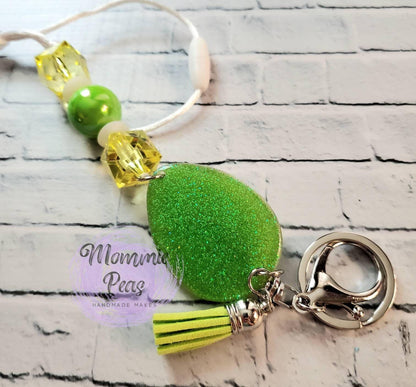 Green glitter teardrop keychain with yellow beads and green tassel on white brick background