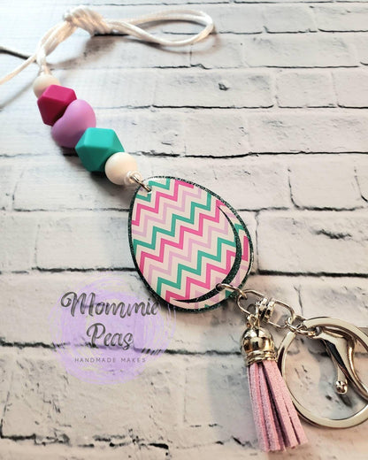 Colorful beaded keychain with chevron patterned teardrop pendant and pink tassel on white brick background