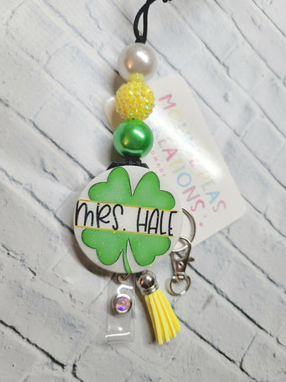 Personalized badge reel with green four-leaf clover, beads, and yellow tassel on white brick background
