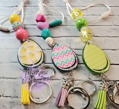 Three handmade Easter egg keychains with colorful beads and tassels on a white brick background