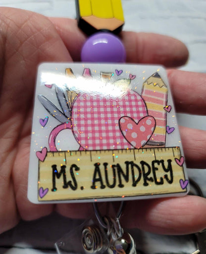 Sparkly badge with pink checkered apple and pencils, labeled Ms. Aundrey, held in hand