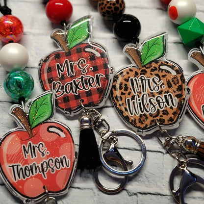 Handmade apple-shaped keychains in red, plaid, and leopard print with teacher names, colorful beads