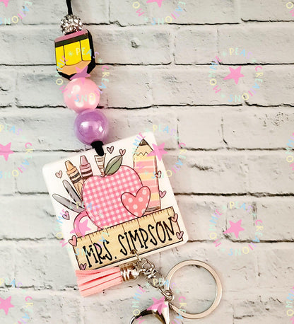 Personalized teacher keychain with apple, crayons, and ruler design labeled Mrs. Simpson on brick background