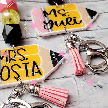 Custom teacher keychains shaped like yellow pencils with names and pink tassels on marble background
