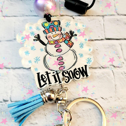 Colorful snowman keychain with blue tassel and text Let It Snow on white brick background