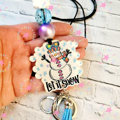 Hand holding snowman keychain with pastel beads and 'Let it snow' text on white brick background