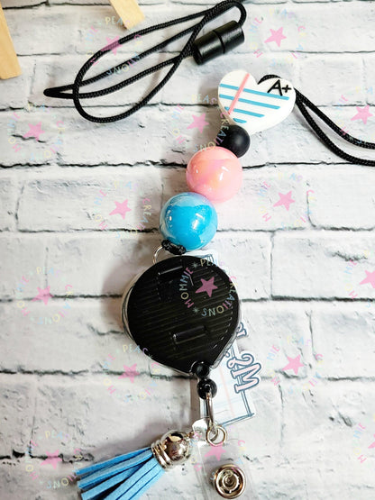 Black retractable badge holder with blue tassel, pastel beads, and heart charm with A+ on white brick background