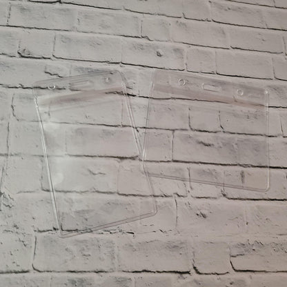 Two clear plastic badge holders on a white brick background for ID cards or passes