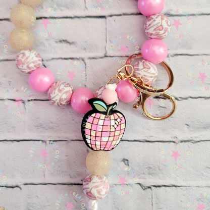 Pink beaded keychain with patterned round beads and apple-shaped charm on a white brick surface