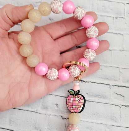 Hand holding pink and beige beaded keychain with apple charm on brick background