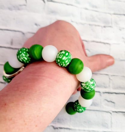 Hand wearing a green and white beaded bracelet with patterned and solid beads against a white brick wall