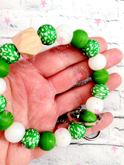 Close-up of a hand holding a green, white, and patterned beaded wrist keychain with a wooden bead and metal ring