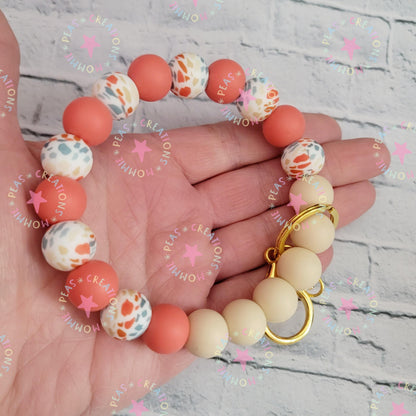 Hand holding beaded bracelet with peach, speckled, and cream beads and gold heart charm on gray stone background