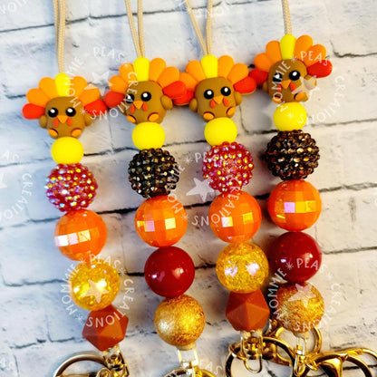 Festive turkey-themed keychains with colorful beads in orange, yellow, brown hues on gray brick background