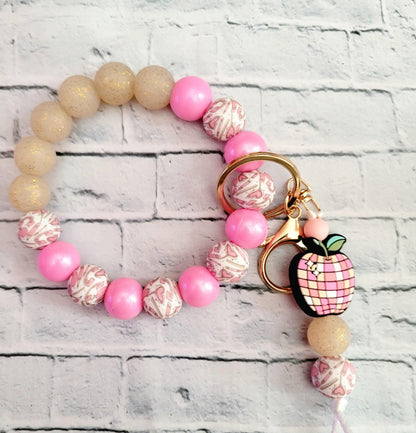 Pink and beige beaded bracelet with heart designs and a pink apple keychain charm on white brick background