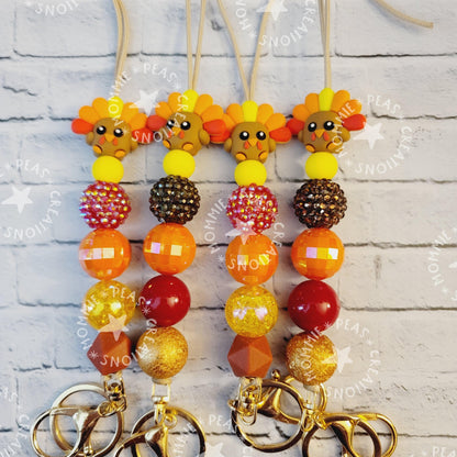 Four turkey-themed beaded keychains with orange, brown, yellow beads on a white brick background