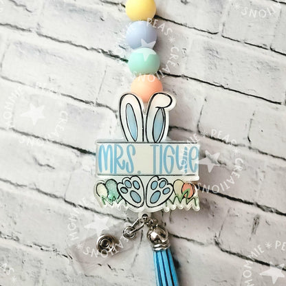 Personalized badge reel with bunny ears, pastel beads, and blue tassel on white brick background