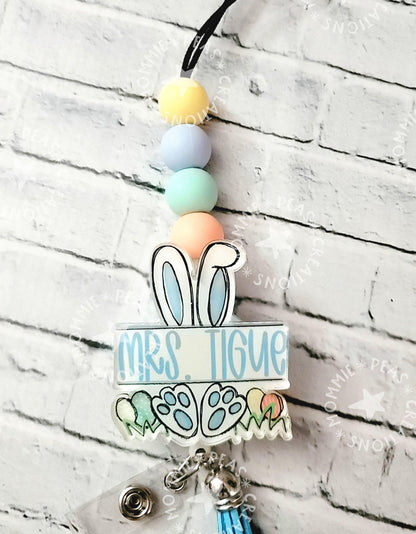 Personalized badge reel with pastel beads, bunny ears, and blue tassel against white brick wall