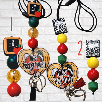 Two personalized teacher keychains with beads, chalkboard and composition book charms against white brick wall