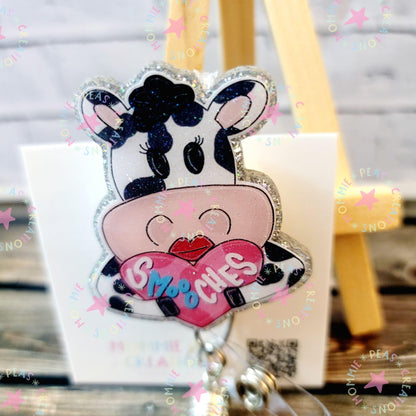 Glittery cow badge holding a pink heart labeled 'SMOOCHES' with a wooden background