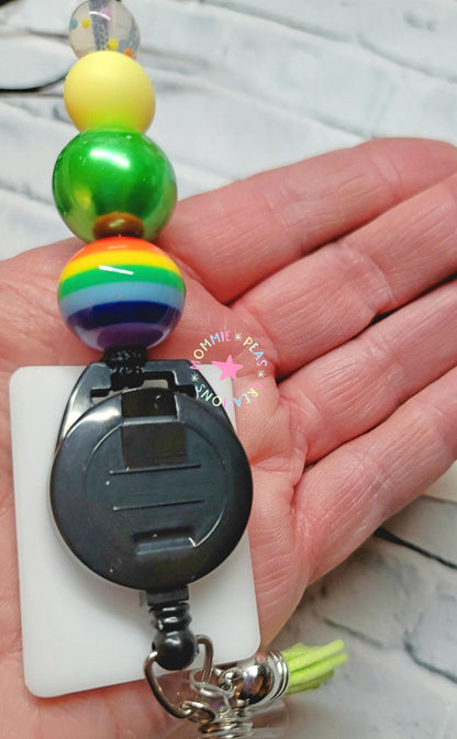 Hand holding rainbow bead badge reel with black retractable clip and tassel keychain
