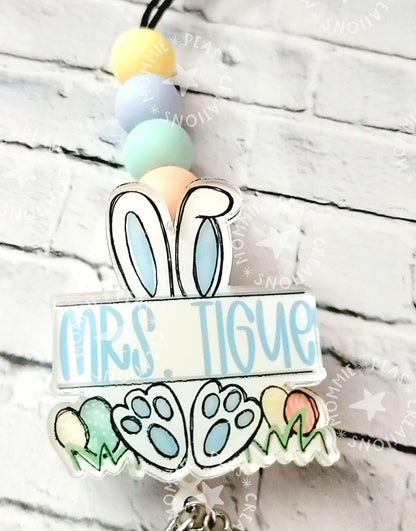 Personalized badge reel with bunny ears, paw prints, pastel beads, and name Mrs. Tigue against white brick wall