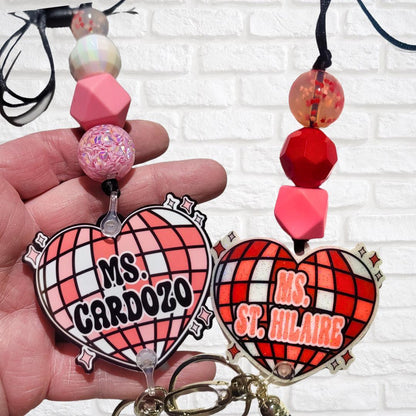 Hand holding two heart-shaped keychains with names Ms. Cardozo and Ms. St. Hilaire, decorated with pink and red beads against white brick background