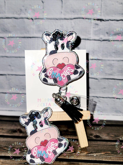 Glittery cow pins with pink heart saying 'Smooches' on wooden surface with white brick background