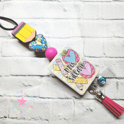 Pink and yellow teacher-themed badge reel with pencil, hearts, and 'Mrs. Silleta' text on white brick background
