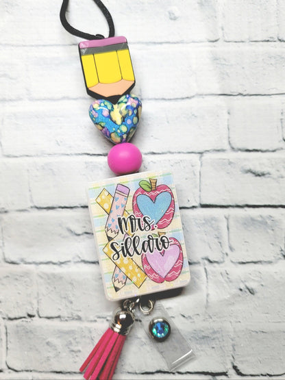 Colorful teacher badge reel with pencil, heart, apples, and pink tassel on brick background