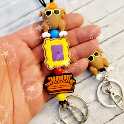 Mommie Peas Creations Fall Turkey Lanyard with Friends-themed charms: a turkey wearing sunglasses, a yellow picture frame, and an orange couch. Features silicone beads and silver hardware.