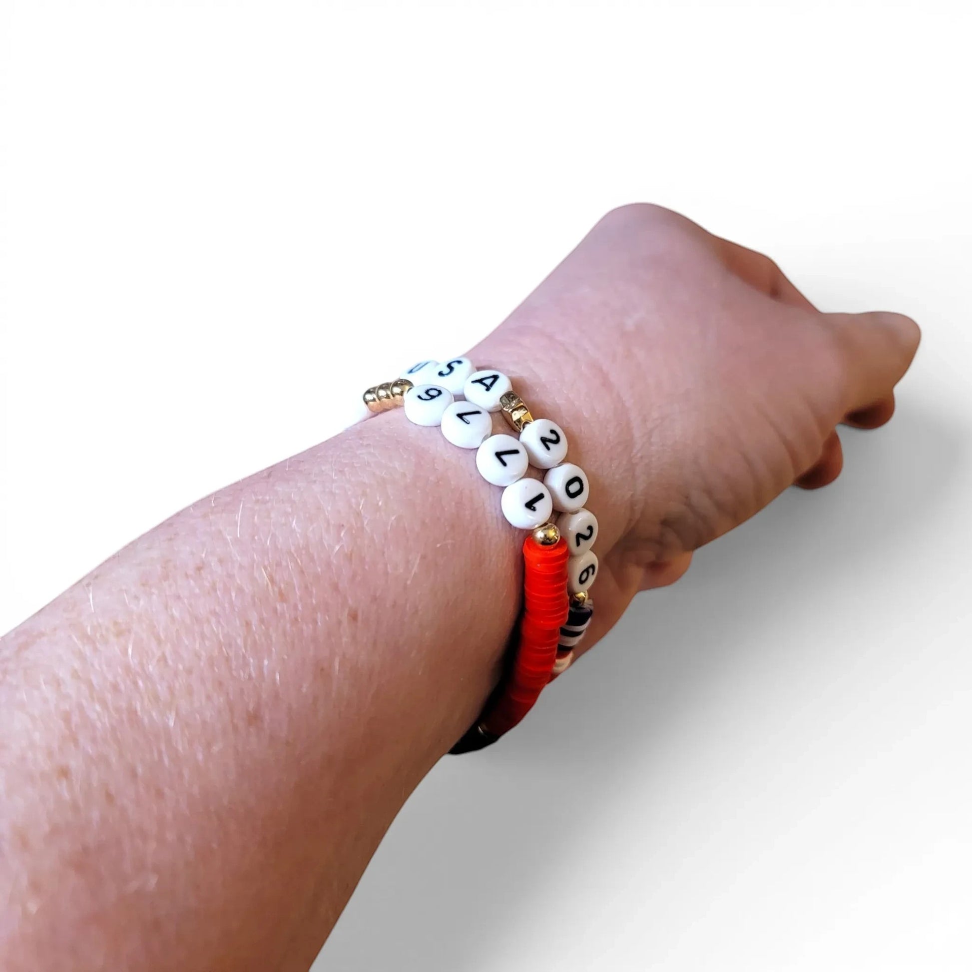 Mommie Peas Creations patriotic beaded bracelets with red vinyl discs, white letter beads spelling USA 2026, and gold star charms.