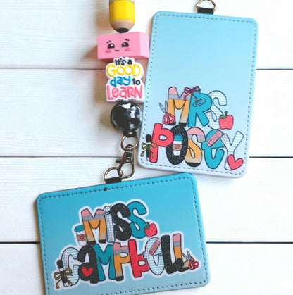 Mommie Peas Creations personalized light blue faux leather ID badge holders with teacher-themed school supply motifs.