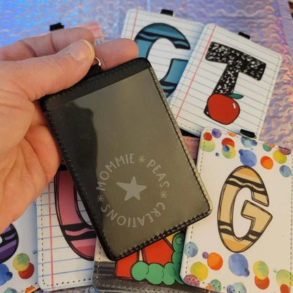 Mommie Peas Creations black PU leather vertical ID badge holder with clear window, featuring fun school-themed designs like letters and crayons on other holders.