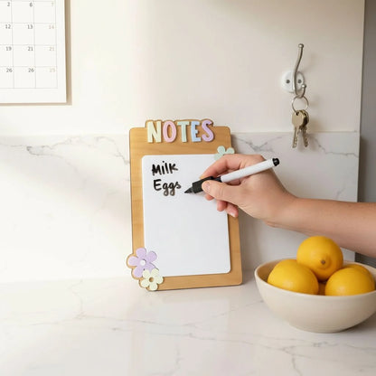 Mommie Peas Creations handmade dry erase notes board with pastel lettering and flower accents. A hand is writing 'Milk Eggs' on the board.