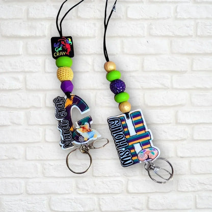 Mardi Gras initial lanyard with purple, green, and gold beads. Features custom acrylic initial charms and a lobster clasp. Mommie Peas Creations.