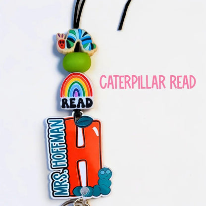 Mommie Peas Creations personalized lanyard with a rainbow, "READ" charm, and an orange "H" with "MRS. HOFFMAN" text, featuring a caterpillar charm.