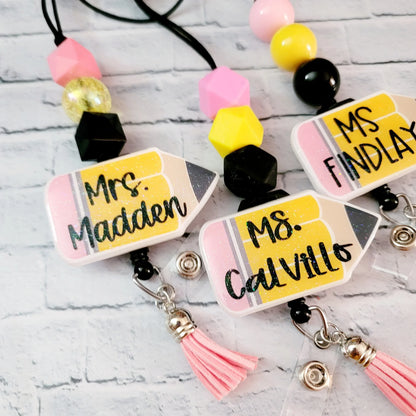 Mommie Peas Creations personalized glitter acrylic teacher lanyard with yellow pencil charm and pink tassel. Features custom names like Mrs. Madden.