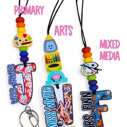 Mommie Peas Creations personalized teacher lanyard with silicone beads, acrylic charm, and custom letter. Features school-themed designs like paint palettes and rulers.